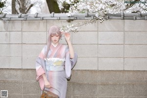 A woman in a pink and gray kimono holding a cherry blossom branch.